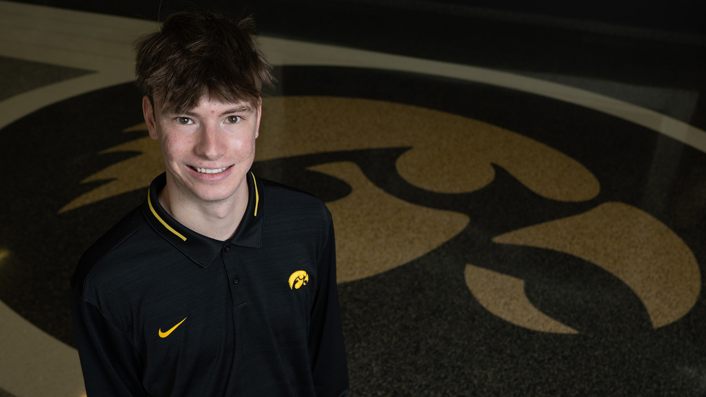 a young man wearing a hawkeye polo stands near a tiger hawk logo that is on the floor at Carver-Hawkeye Arena