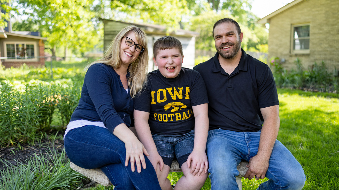 A boy and his parents sitting on a bench outside smiling